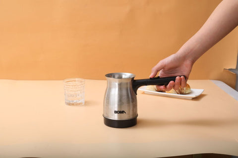 Person using a Bialetti coffee maker on a beige surface with a glass of water and a plate of food in the background.