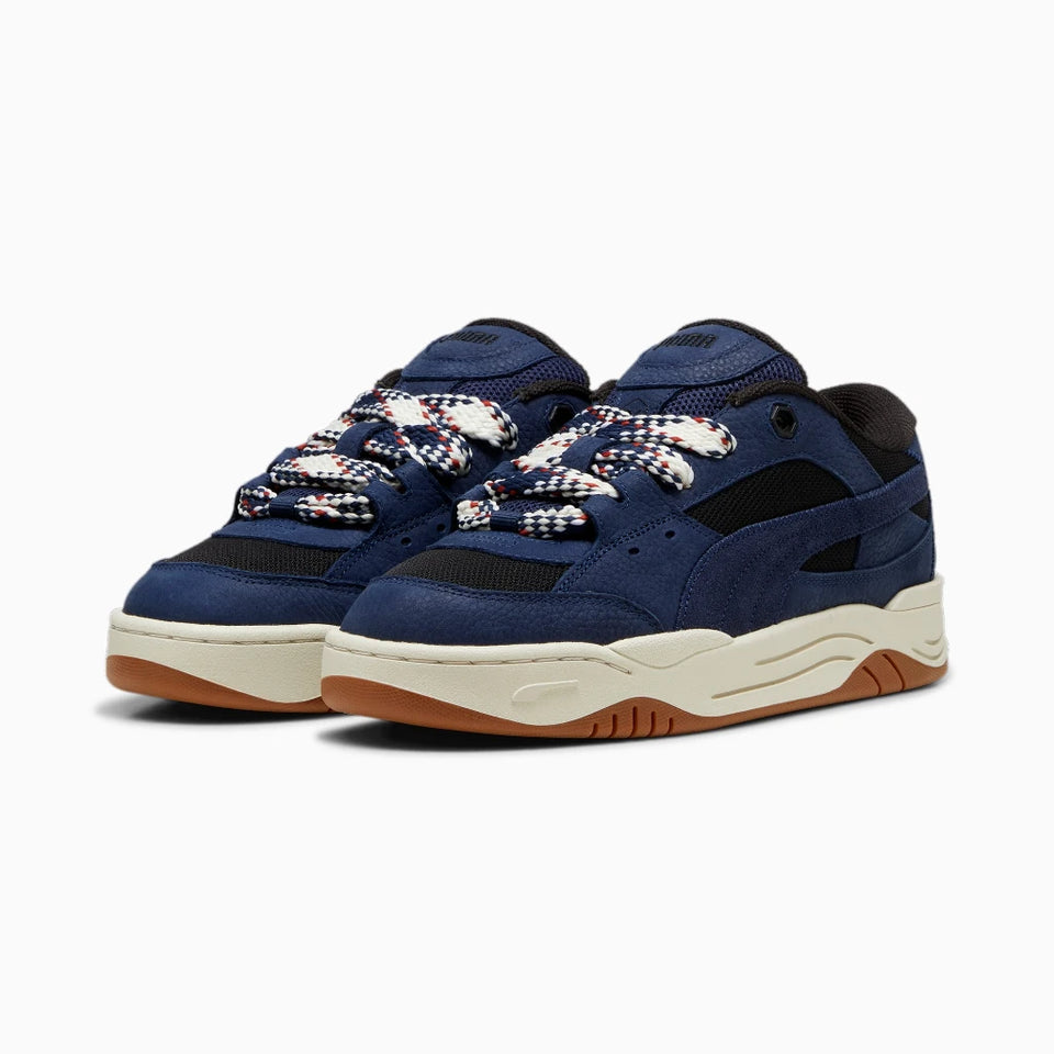 A pair of Puma sneakers in navy blue suede and mesh, featuring patterned white, red, and blue laces, a cream midsole, and a brown gum outsole, shown against a plain white background.