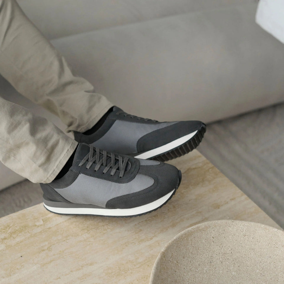A person wearing gray and black athletic sneakers with beige pants, sitting on a light beige sofa and resting their feet on a light wood table next to a textured ceramic bowl.