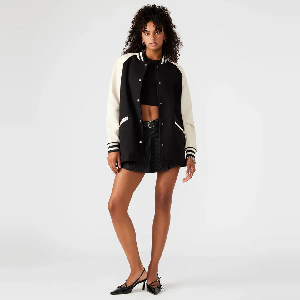 A model wearing a black and off-white varsity jacket, black crop top, black denim shorts, and black pointed-toe heels, standing against a light gray studio background.