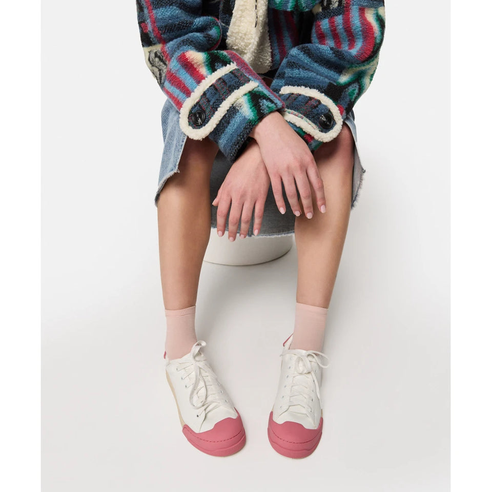 A person seated on a white stool wearing a multicolored striped fuzzy cardigan, denim skirt, pink socks, and cream low-top sneakers with pink rubber toe caps, against a light gray studio background.