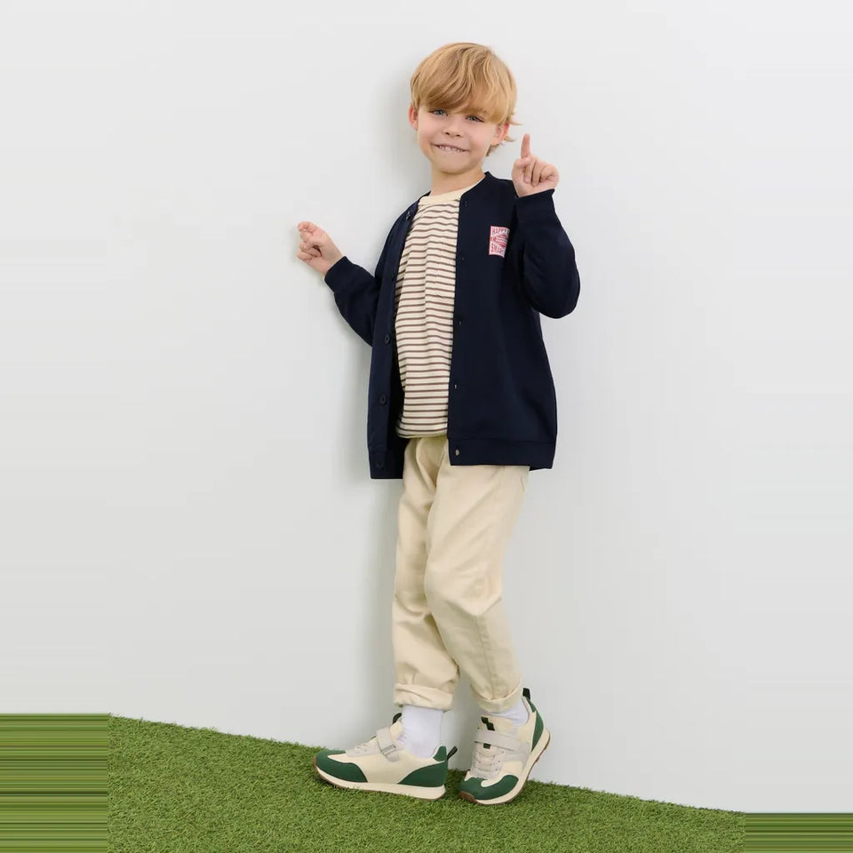 A young boy stands against a white wall wearing a navy blue zip-up cardigan over a beige and white striped shirt, paired with beige pants and green-and-white sneakers.