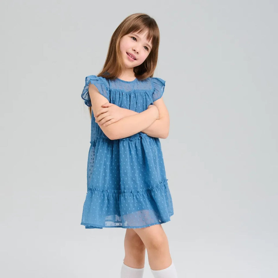A young girl with light brown hair wearing a tiered blue eyelet dress with white socks, standing against a light gray studio background.
