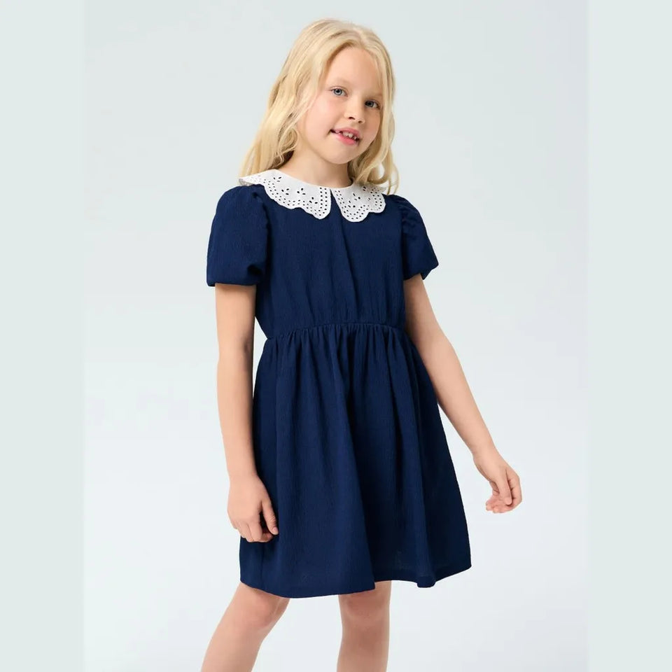 A young blonde girl wearing a navy blue short-sleeve dress with a white eyelet Peter Pan collar, standing against a light gray studio background.