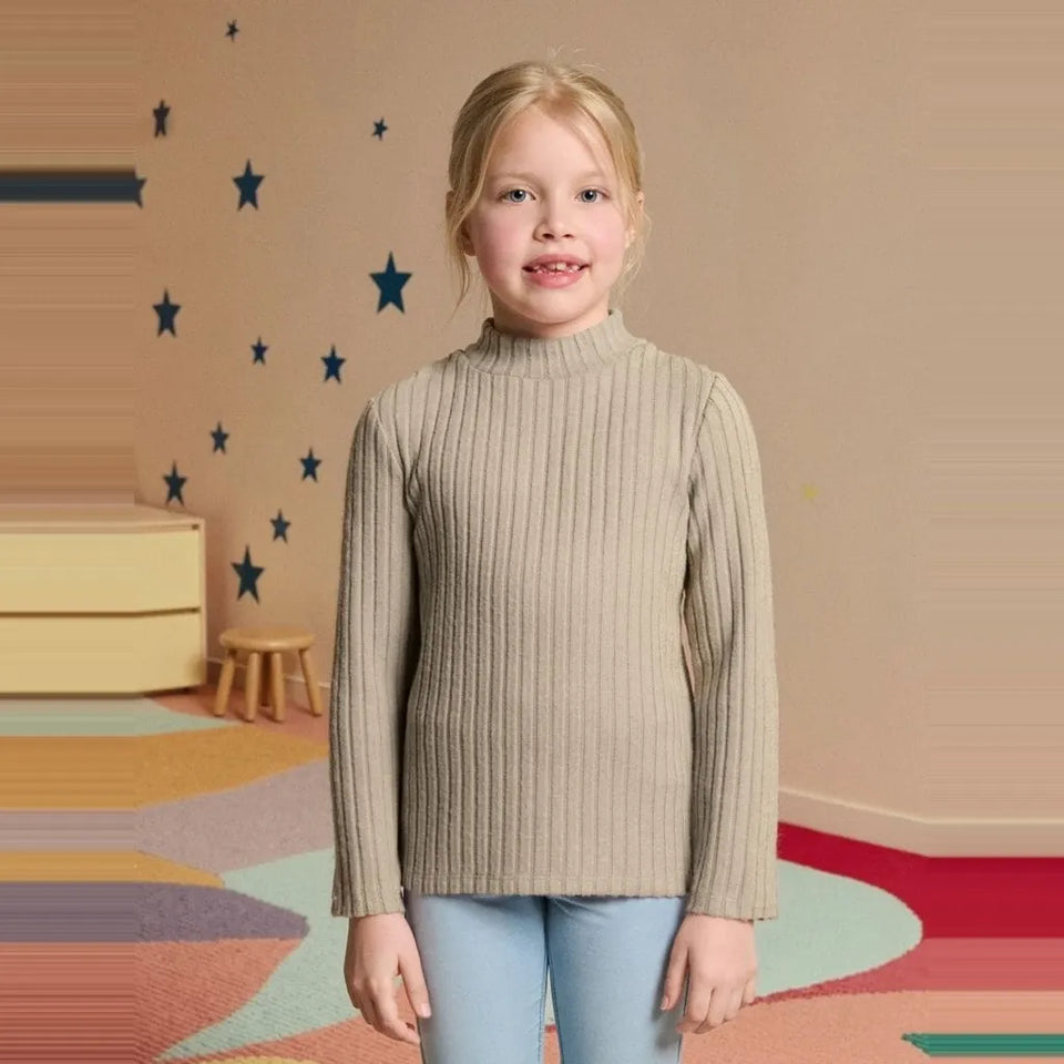 A young girl with blonde hair stands in a child's bedroom wearing a beige ribbed mock neck sweater and light blue jeans.