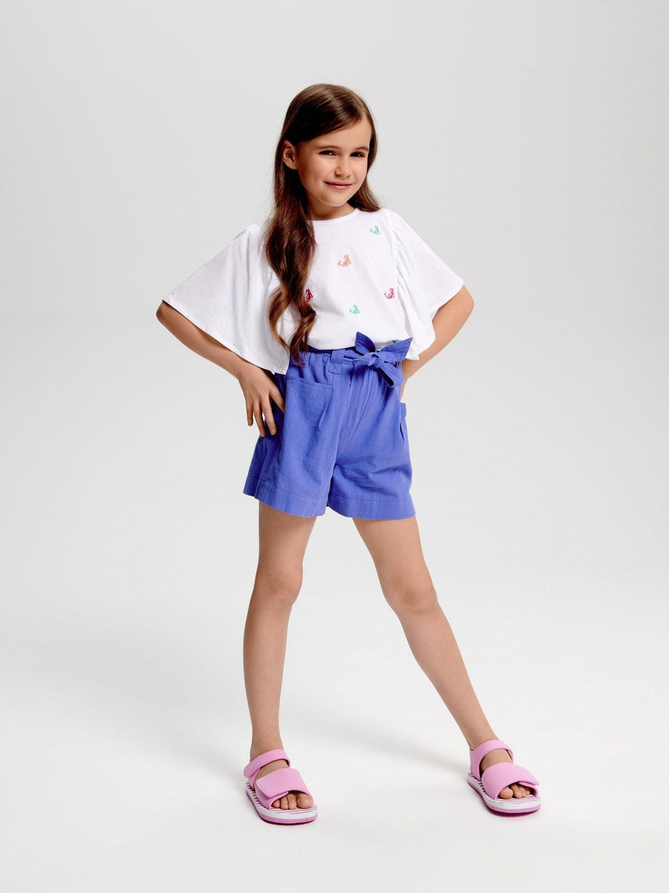 Young girl wearing a white t-shirt with colorful designs and blue shorts on a light gray background