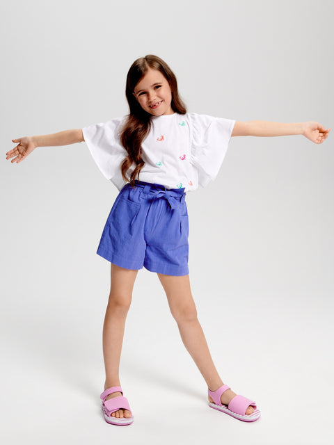 Young girl wearing a white t-shirt with colorful designs, blue shorts, and pink sandals on a light gray background