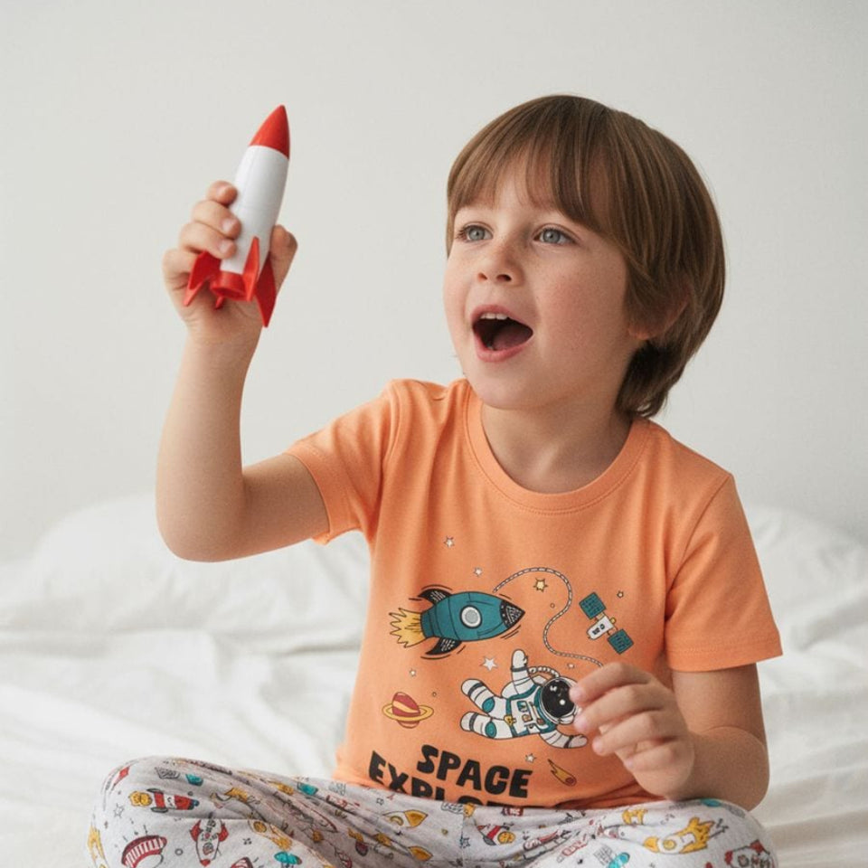 A young child sitting on a white bed wearing an orange t-shirt and patterned pajama pants, holding a red and white toy rocket up in the air with a surprised expression.
