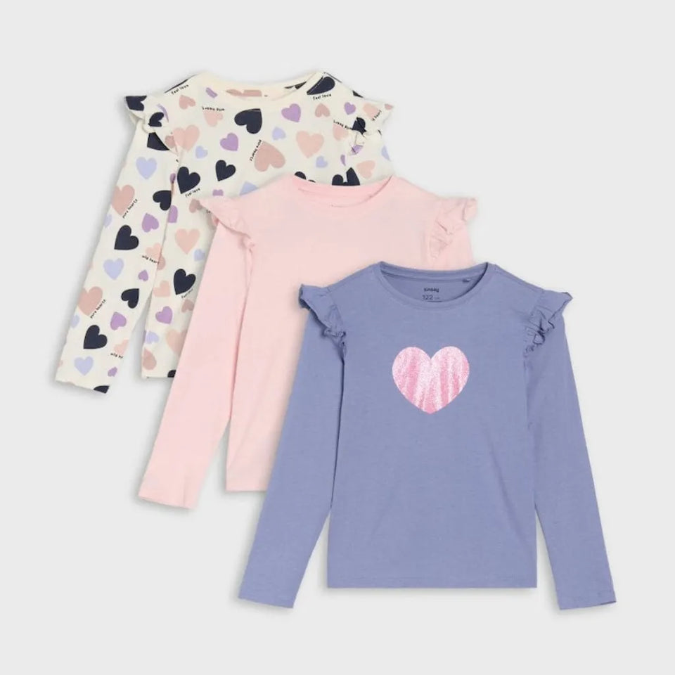 Three girls' long-sleeve ruffle-shoulder tops arranged in an overlapping flat lay on a light gray background. The top in back is cream with scattered black, pink, and purple hearts; the middle is light pink; the front is lavender with a pink heart graphic on the chest.