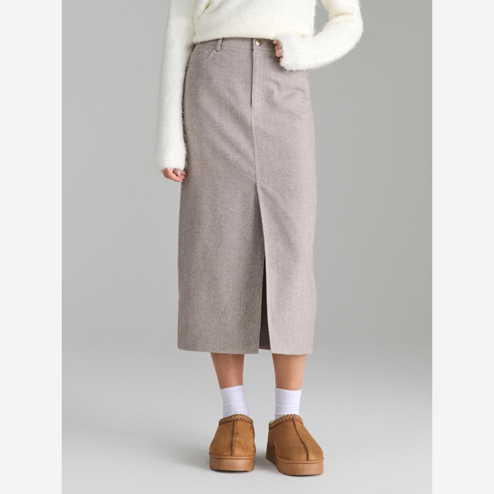 A model wearing a mid-calf, knee-length taupe knit skirt with a front slit, styled with a white fuzzy sweater, white socks, and brown suede slip-on shoes against a neutral gray background.