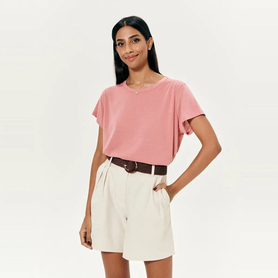 A woman modeling a dusty pink short-sleeve t-shirt tucked into cream-colored pleated shorts with a brown belt, against a plain white studio background.