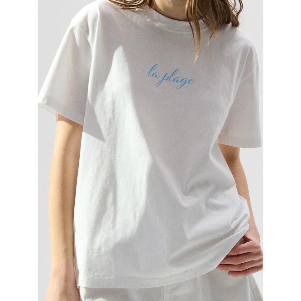 A person wearing an oversized white short-sleeve T-shirt with the words 'la plage' printed in light blue script across the chest, shown against a plain light gray background.