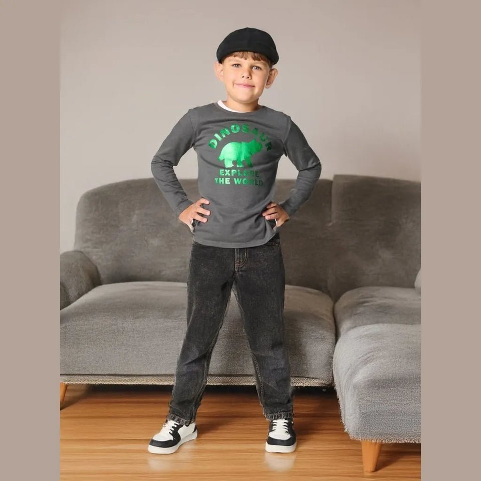 A young boy stands in front of a gray sectional sofa wearing a dark gray long-sleeve t-shirt with a green dinosaur graphic, dark gray jeans, a black cap, and sneakers.