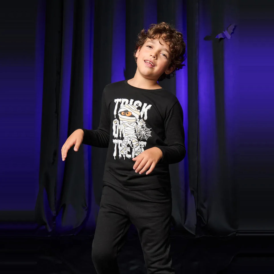 A young boy modeling a black long-sleeve t-shirt with a white 'Trick or Treat' mummy graphic, posed against a dark curtain backdrop with blue lighting.