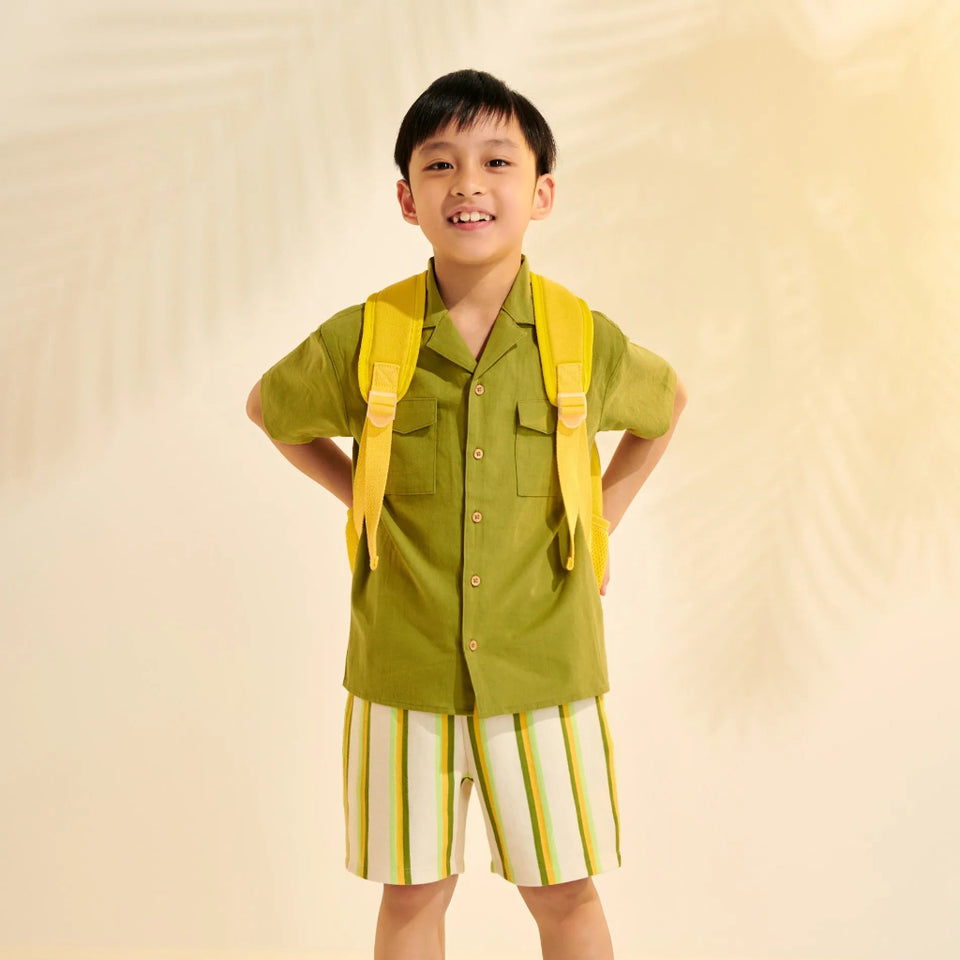 A young boy stands with hands on hips wearing a matching olive green short-sleeve button-up shirt and striped shorts, styled with a bright yellow backpack and green sandals against a warm cream backdrop.