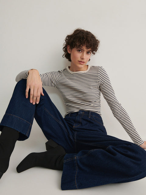 Person wearing a striped shirt and wide-leg jeans sitting on the floor against a plain background