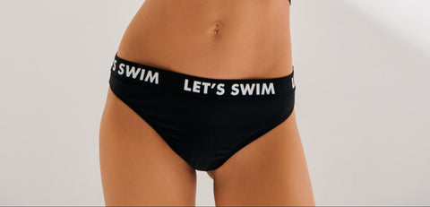 Woman wearing a black 'Let's Swim' bikini set with sunglasses on a white background