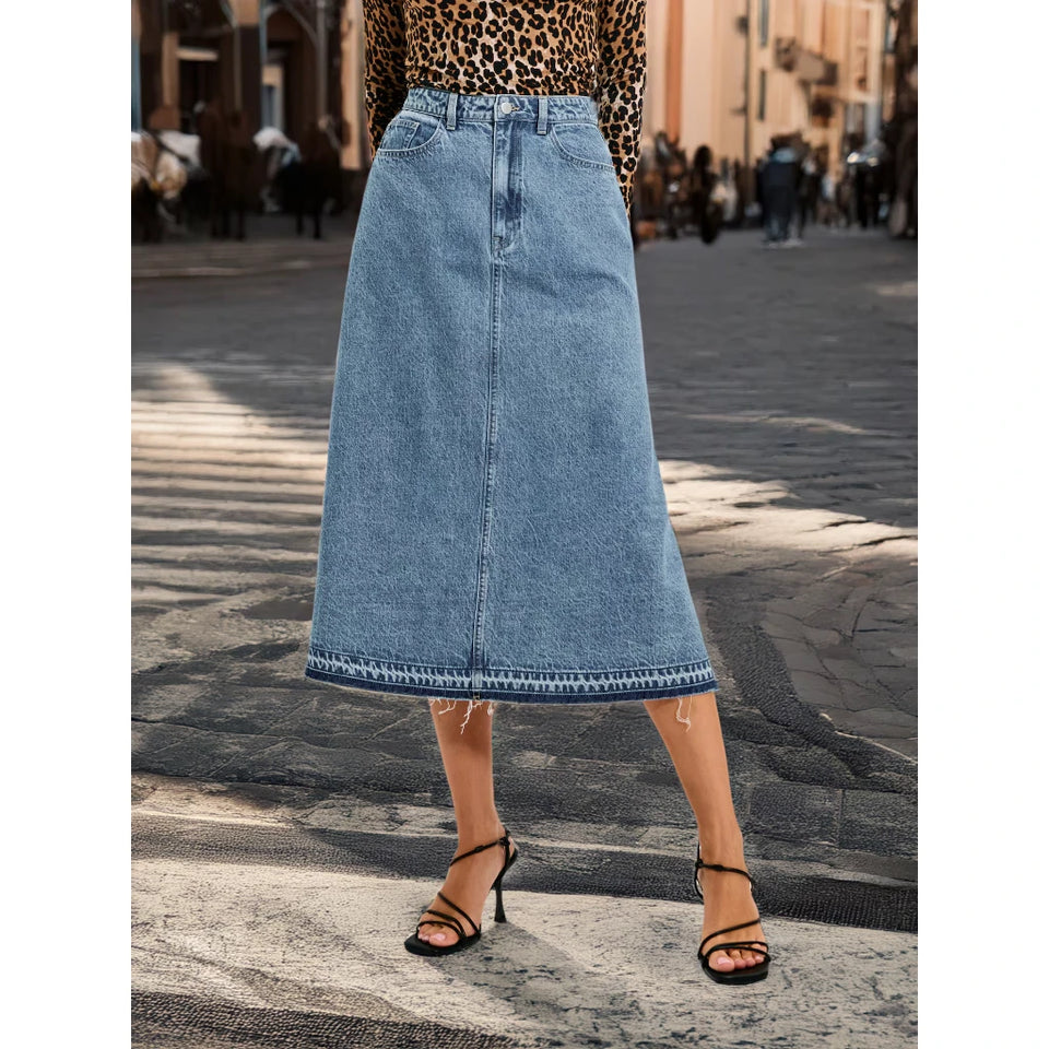 A woman wearing a light blue denim midi skirt with a raw hem, paired with a leopard print top and black strappy heels, standing on a cobblestone street.