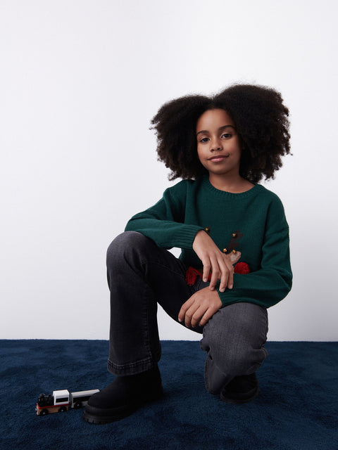 Child wearing a green sweater with red buttons sitting on a blue surface against a white background