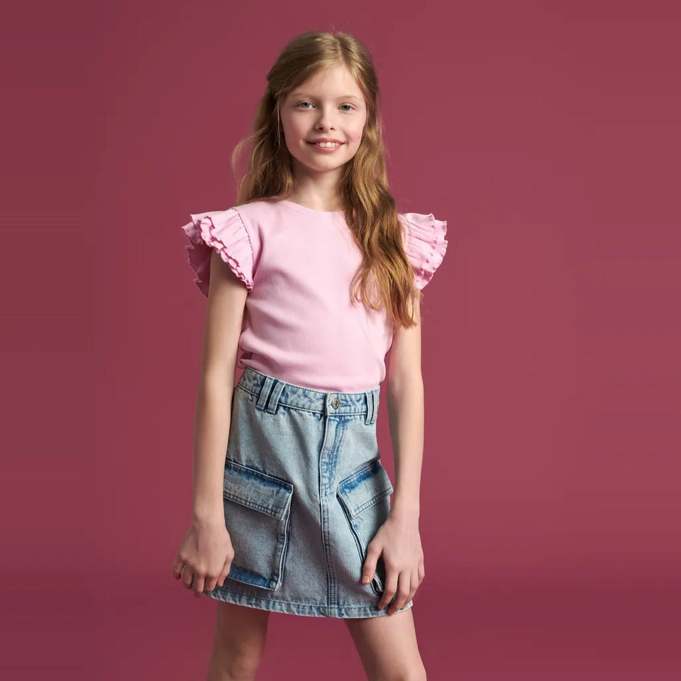 A young girl with long light brown hair stands against a solid dusty rose background, wearing a light pink short-sleeve top with ruffled flutter sleeves and a light blue denim cargo mini skirt.
