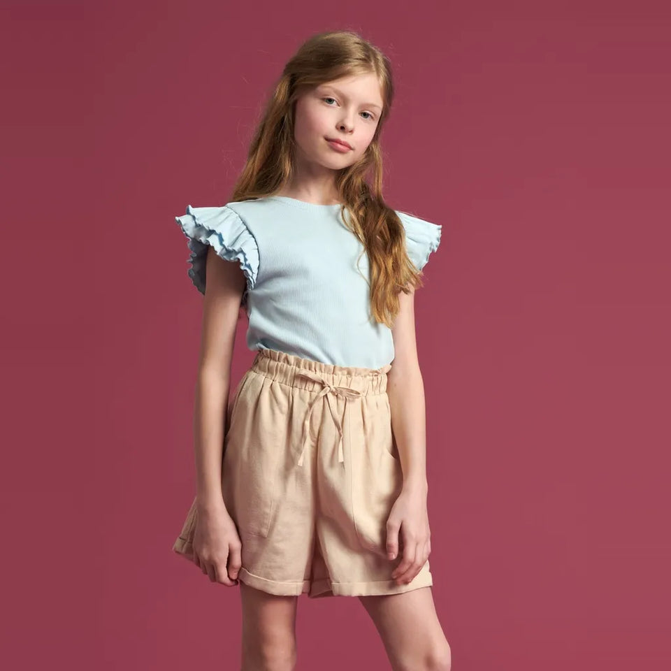 A young girl with long light brown hair stands against a solid dusty rose background, wearing a light blue ruffled-sleeve top and beige linen shorts.