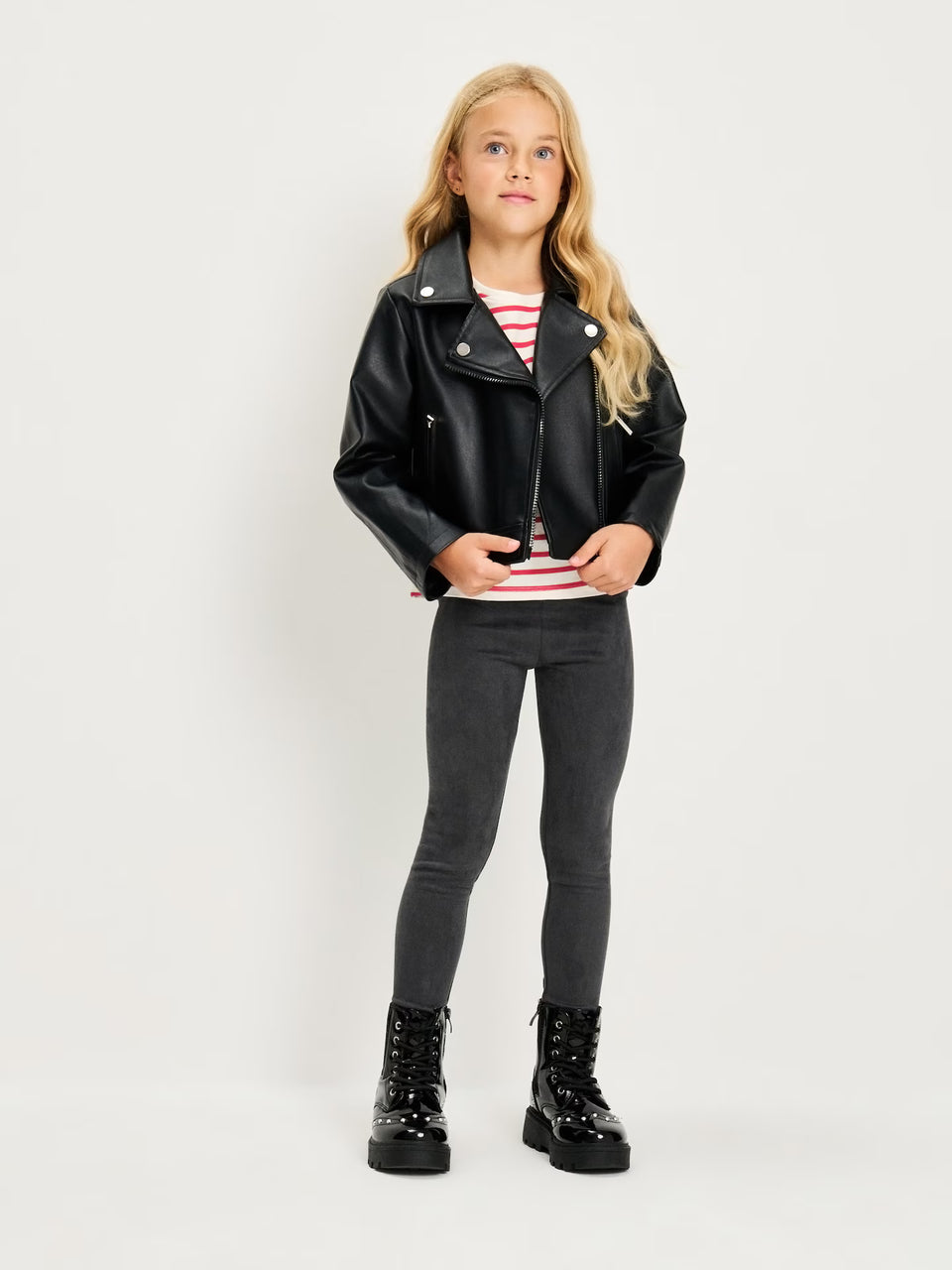 A young girl stands against a light gray studio background wearing a black faux-leather moto jacket, a red and white striped long-sleeve shirt, dark gray leggings, and black lace-up boots.