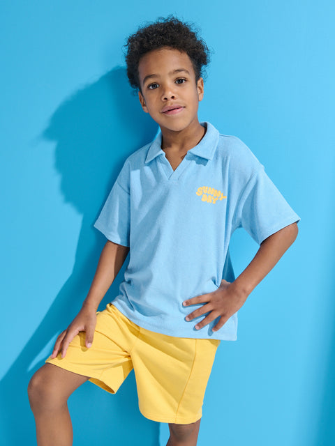 Child wearing a light blue polo shirt and yellow shorts against a blue background