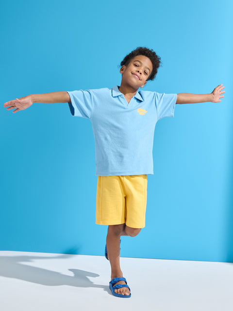 Child wearing a light blue shirt and yellow shorts against a blue background