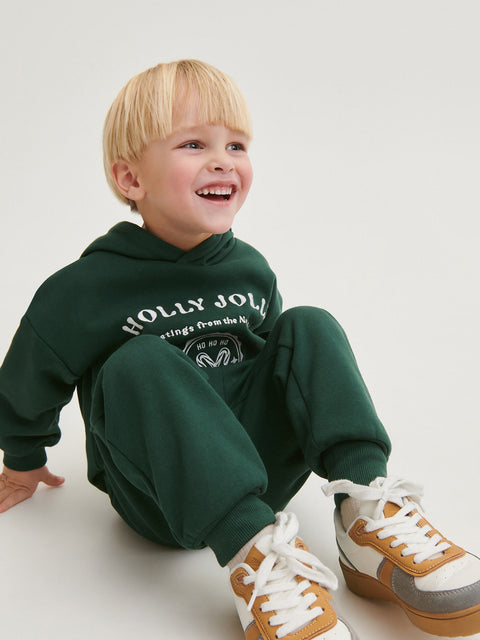 Child wearing a green hoodie and pants with visible branding, sitting on a white floor.