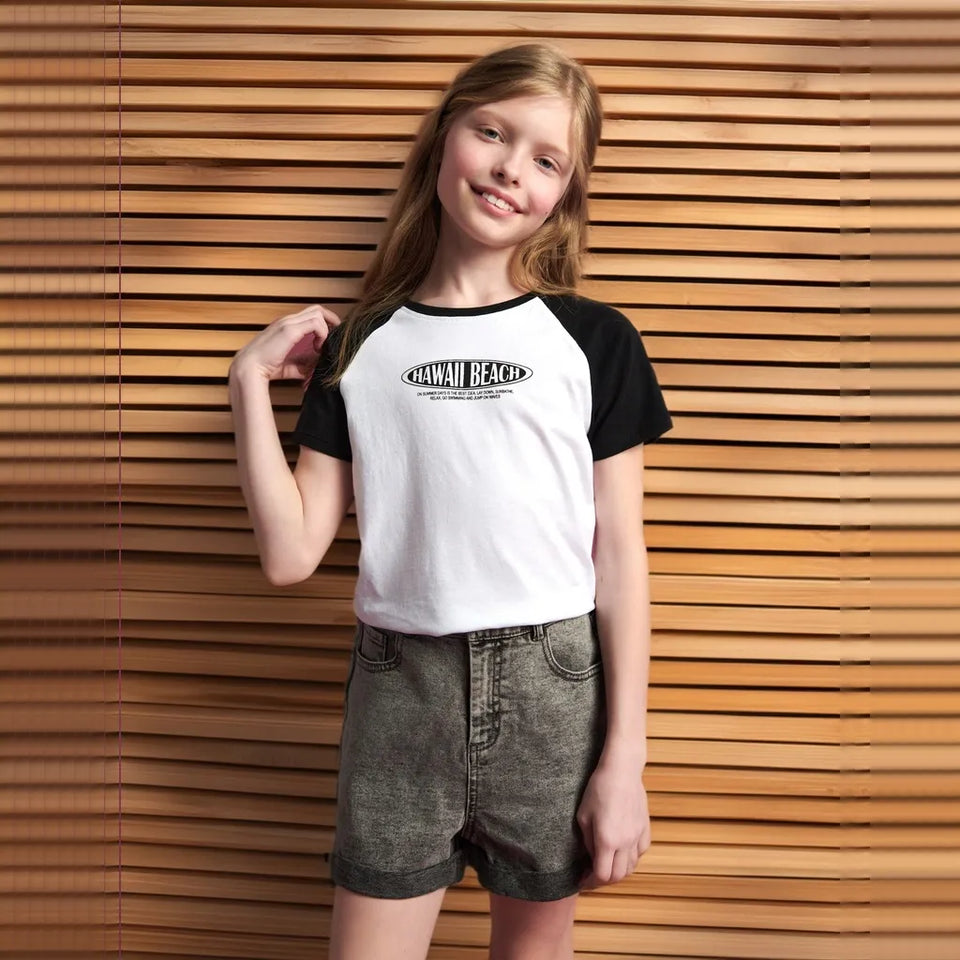 A young girl with long light brown hair stands in front of a wooden slat wall, wearing a raglan-style short-sleeve t-shirt with a white body and black sleeves, featuring a black graphic that reads 'HAWAIIAN BEACH'. She is also wearing gray denim shorts.