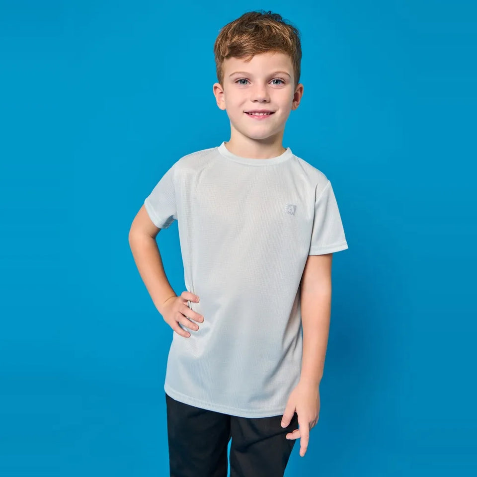 A young boy with short brown hair stands against a solid blue studio background, wearing a light gray short-sleeve crewneck t-shirt and black pants.