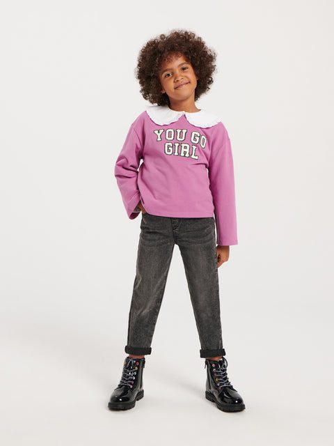Child wearing a pink sweatshirt with text, black pants, and black boots on a white background