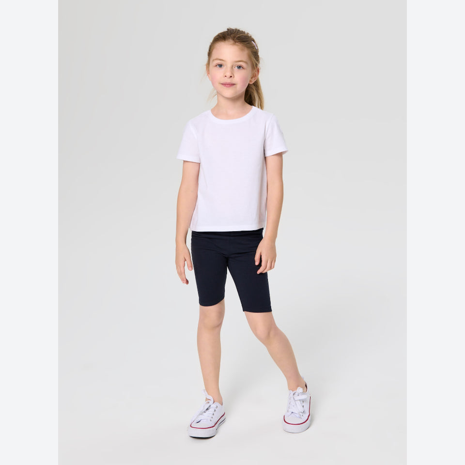 A young girl modeling a white short-sleeve crewneck t-shirt, dark navy bike shorts, and white low-top sneakers with red stripes, standing against a light gray studio background.
