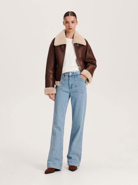 Person wearing a brown shearling jacket, white shirt, and blue jeans on a light gray background