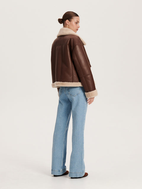 Woman wearing a brown leather jacket with fur trim and blue jeans on a white background