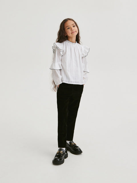 Person wearing a white blouse with ruffled sleeves and black pants on a light gray background