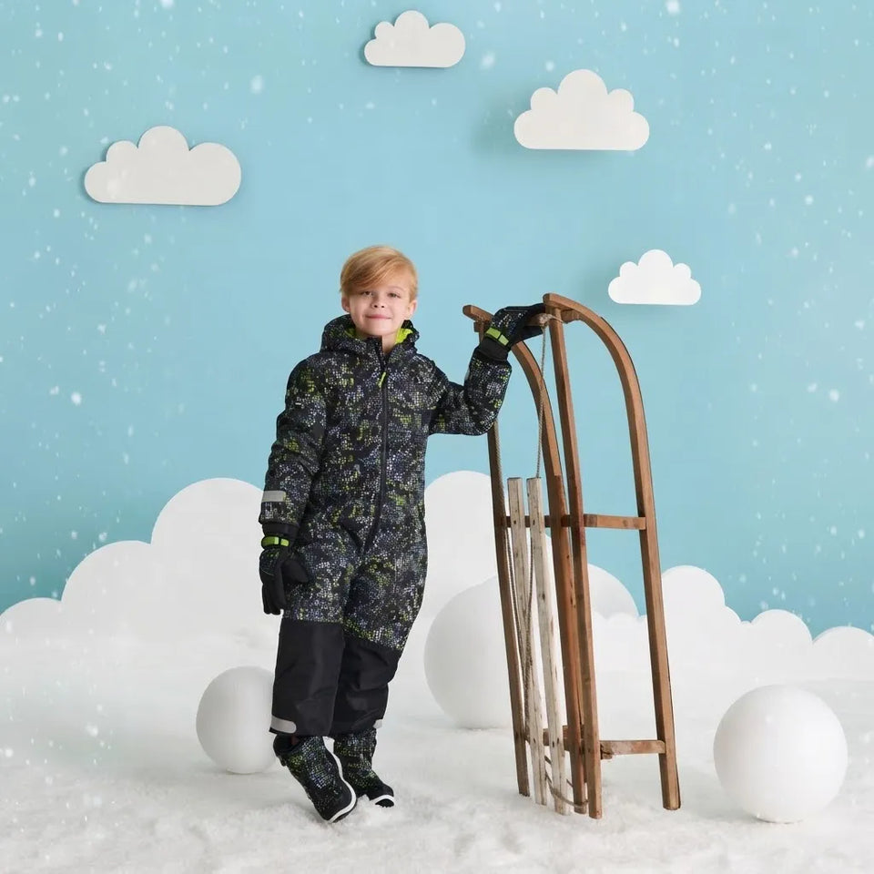 A young boy wearing a dark camouflage winter jacket and black snow pants stands in a snowy studio setup next to a wooden sled. The background features a light blue sky with white clouds and falling snow effects.