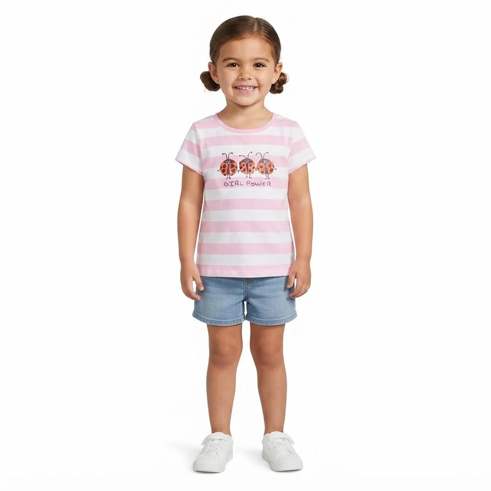 A young girl with brown pigtails stands against a white background wearing a pink and white striped t-shirt with ladybugs and the text 'LADYBUGS', blue denim shorts, and white sneakers.