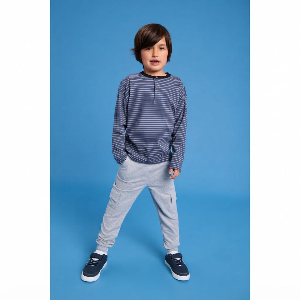 A young boy modeling a long-sleeve striped henley shirt in navy and light gray, paired with light gray jogger pants and navy sneakers, standing against a solid blue studio background.