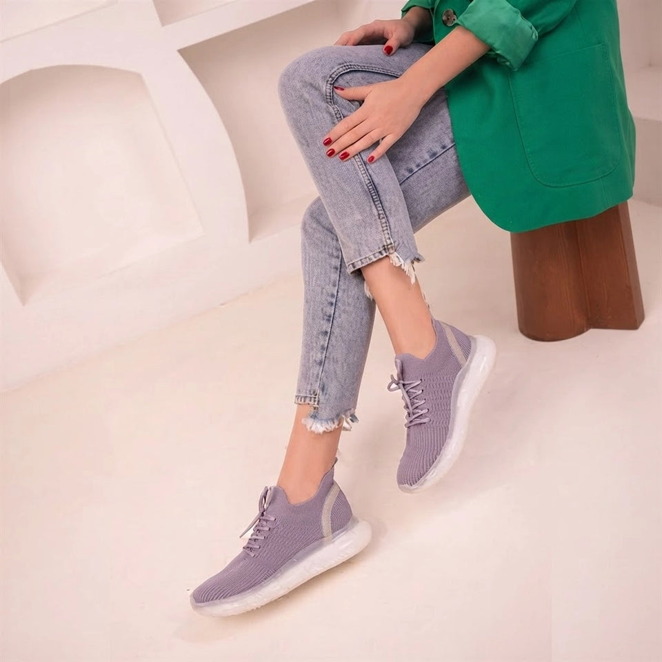 A person seated on a wooden stool wearing light purple knit slip-on sneakers with white soles, paired with light blue jeans and a green top in a bright indoor setting.