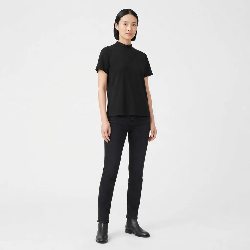A model wearing a black short-sleeve mock neck t-shirt, black skinny jeans, and black leather ankle boots, standing against a solid light gray background.