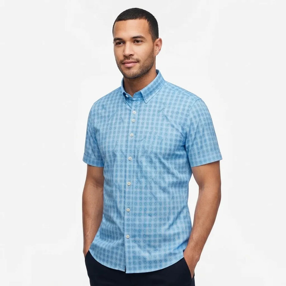 A man wearing a light blue short-sleeve button-down shirt with a subtle geometric pattern, paired with dark pants, standing against a plain white studio background.
