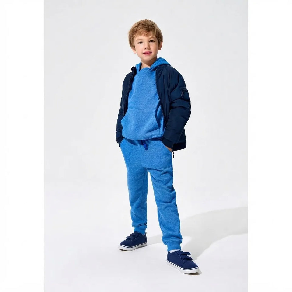 A young boy modeling a blue children's tracksuit consisting of a heather blue hoodie, matching blue sweatpants, and navy blue slip-on shoes, styled with a navy blue puffer jacket over his shoulders, against a plain light gray studio background.
