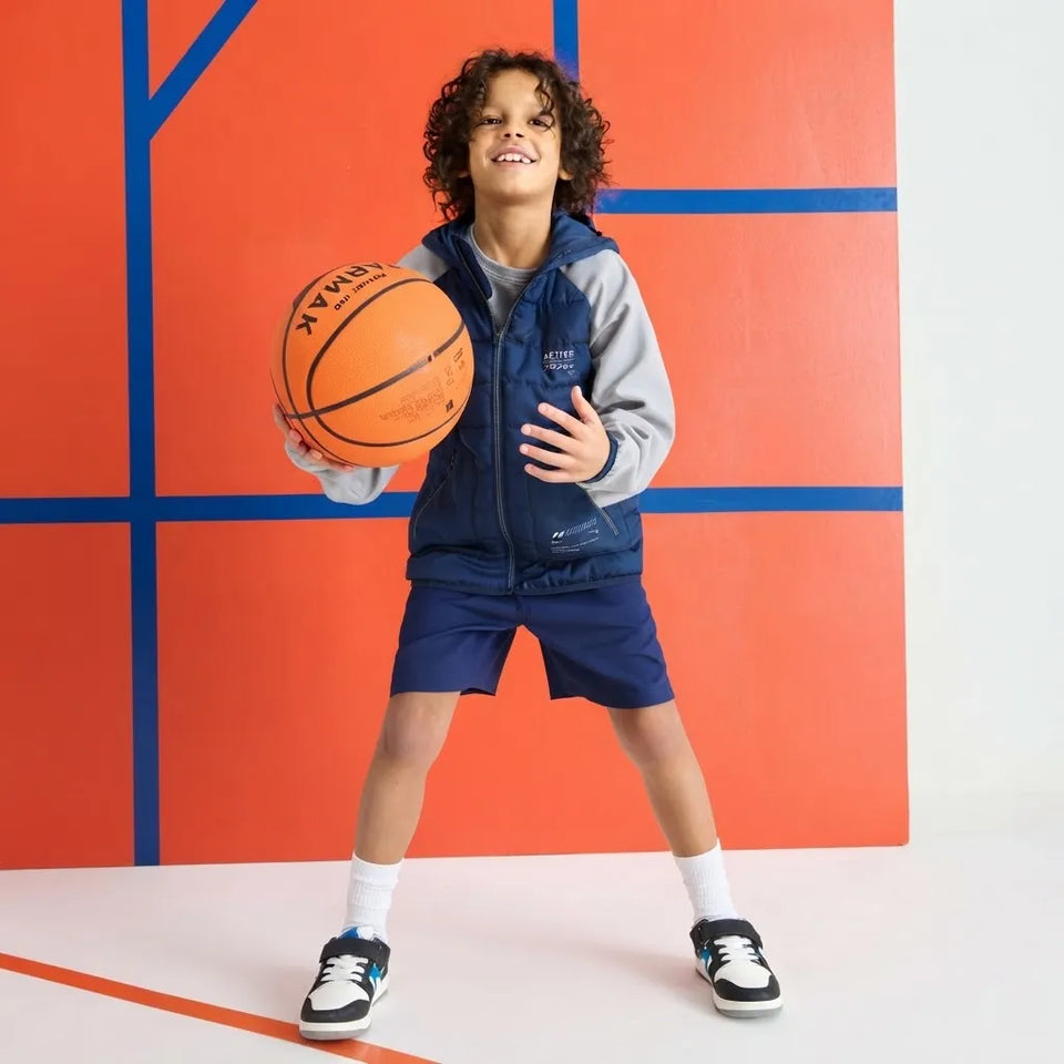 A young boy with curly hair stands in a studio wearing a navy blue and gray zip-up athletic jacket, matching navy shorts, white socks, and black sneakers, while holding an orange basketball. The background is a bold orange wall with blue geometric lines.