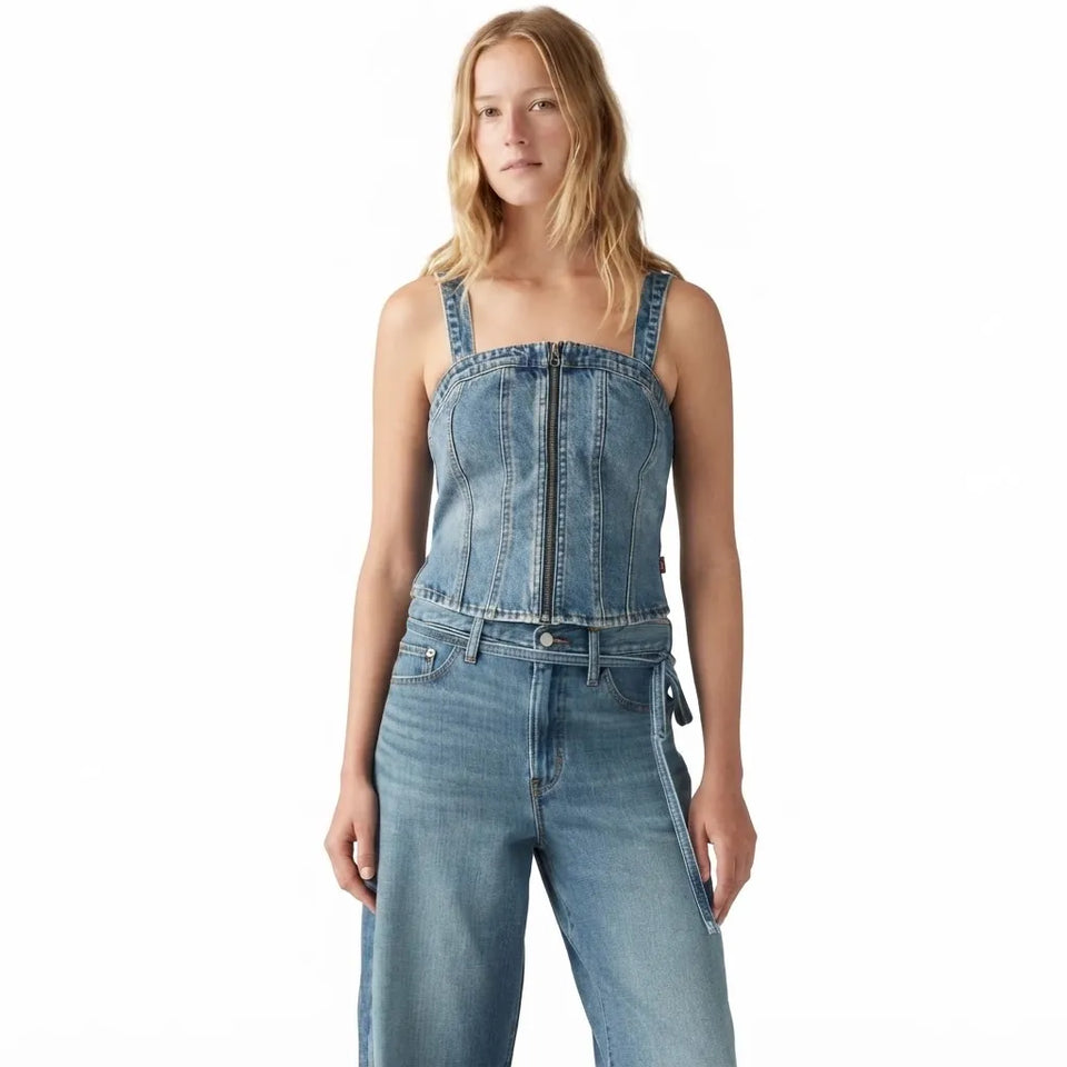 A woman modeling a light blue denim jumpsuit with a square neckline, front zipper, and wide-leg jeans on a plain white background.