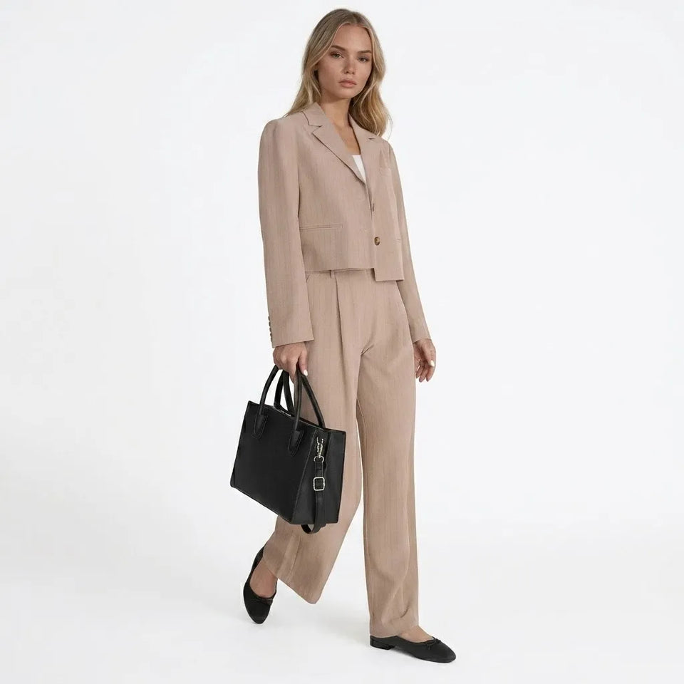 A woman models a beige tailored two-piece suit consisting of a cropped blazer and matching wide-leg trousers, paired with black flats and holding a black structured handbag against a plain white studio background.