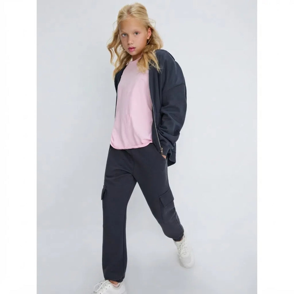 A young girl modeling a dark grey zip-up jacket over a light pink t-shirt, paired with dark navy cargo pants and white sneakers, against a light gray studio background.