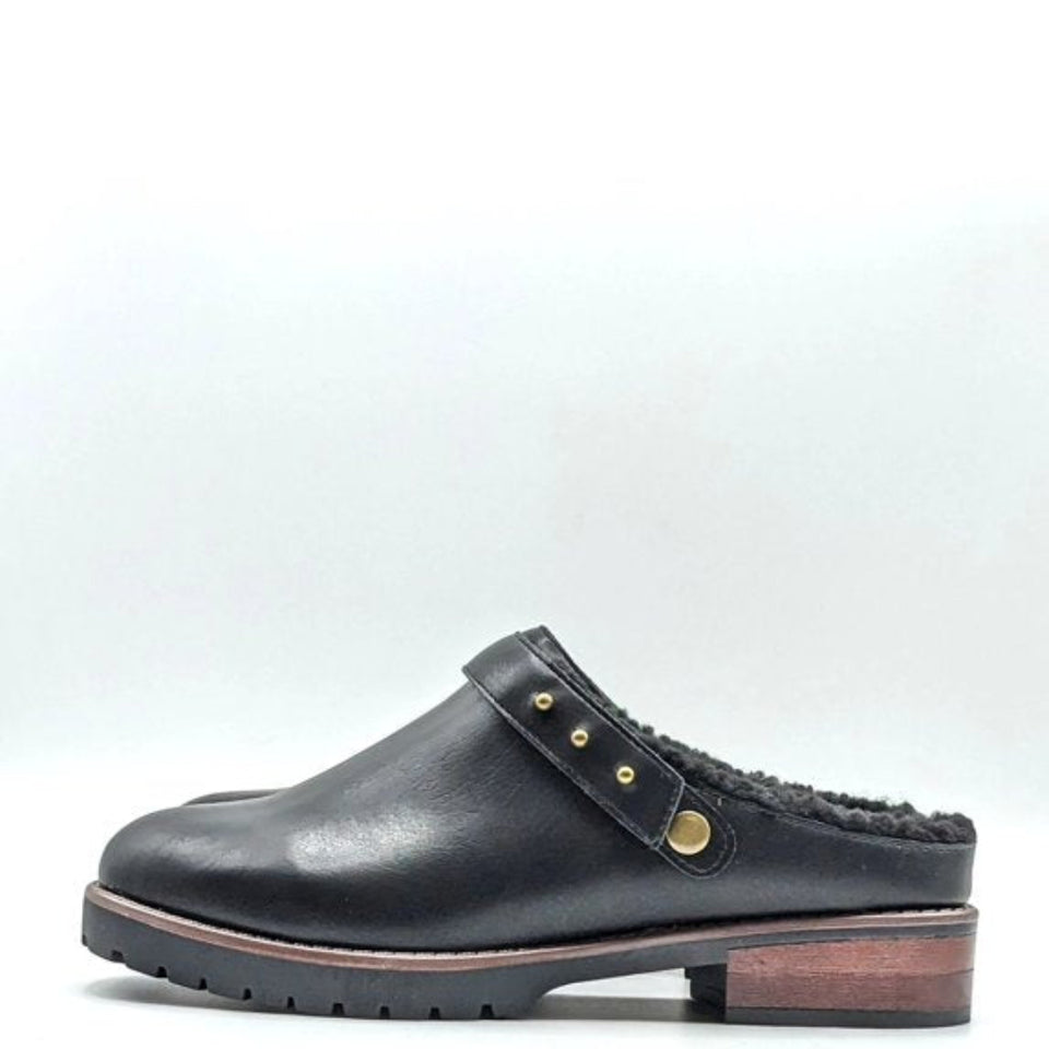 A black leather clog-style slipper with a fuzzy black shearling lining, a strap adorned with gold-tone studs, and a low stacked heel, shown against a plain light gray background.