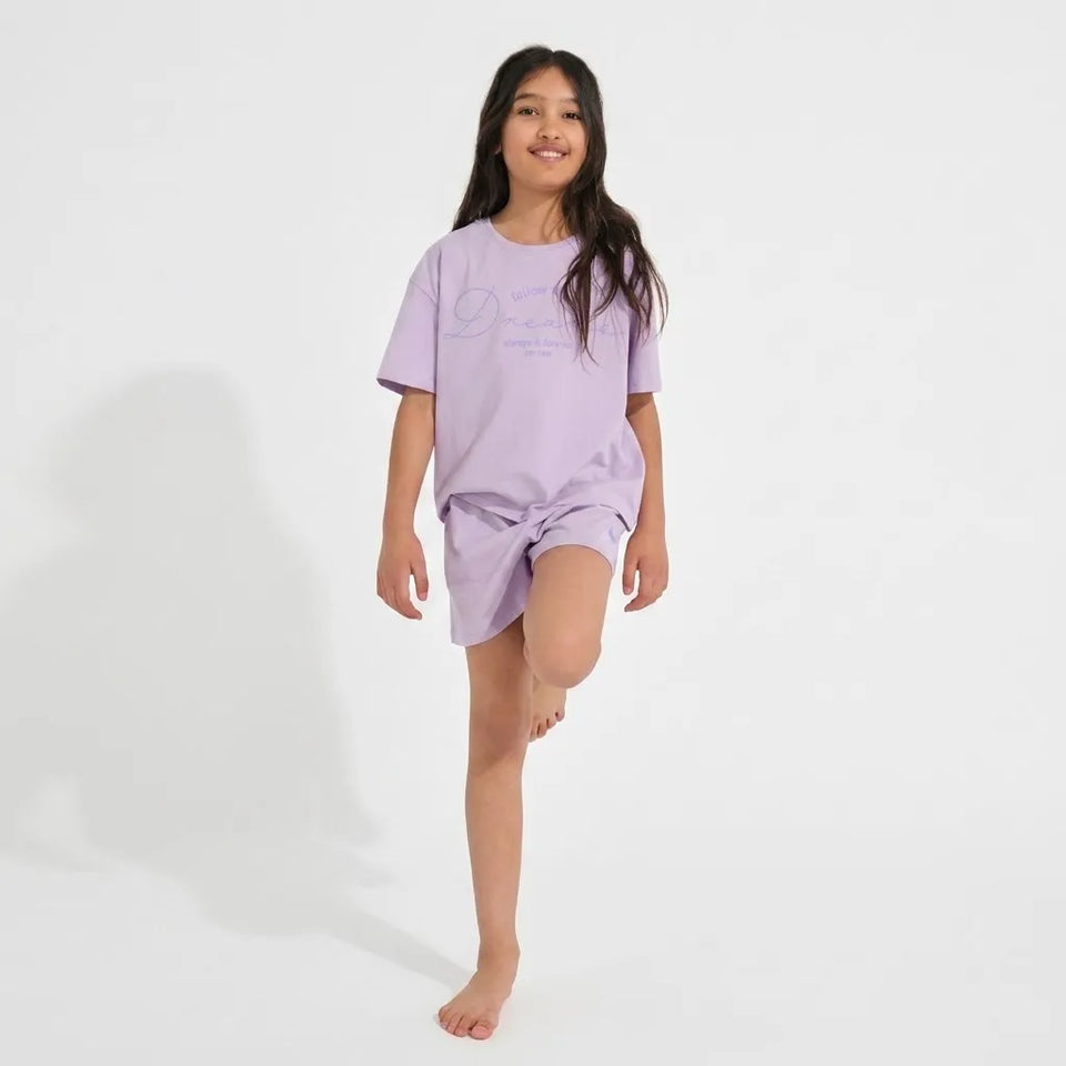 A young girl modeling a light purple short-sleeve pajama set with a matching graphic print, standing barefoot against a plain white studio background.