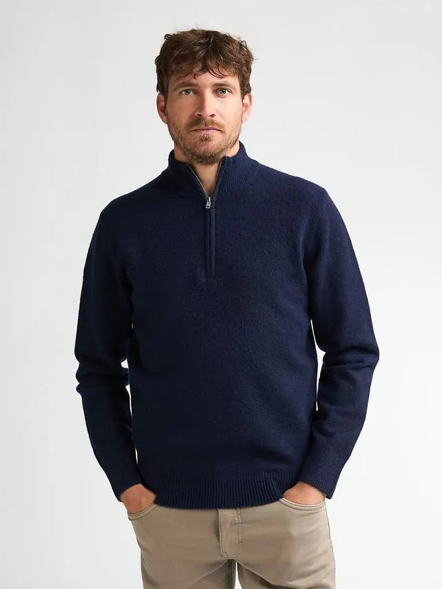 A man wearing a navy blue quarter-zip knit sweater and beige trousers, standing against a plain light gray studio background.